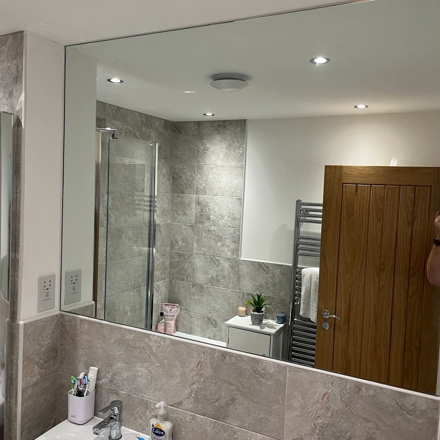 Wide frameless mirror reflecting a grey tiled bathroom and shower